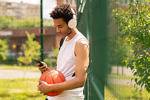 Young handsome athlete in sportswear listening to music in headphones and scroling in smartphone after game