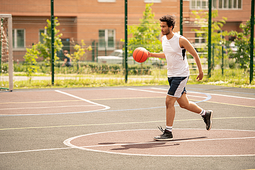 Young basketballer in sportswear with ball running down playground while training on summer day