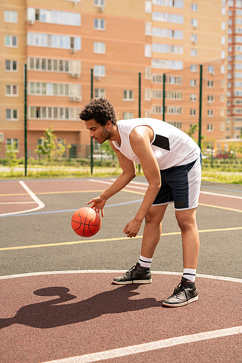 Young basketball player practicing exercise with ball while bending forwards over court in urban environment