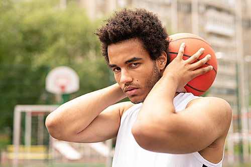 Young serious sportsman holding ball for playing basketball behind neck while standing in front of camera
