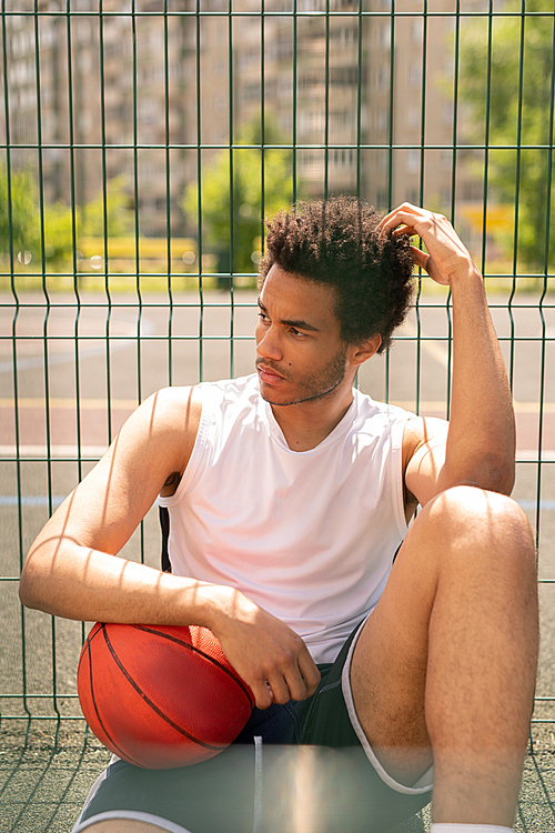 Young serious restful basketball player with ball sitting by fence of playground while having rest after training
