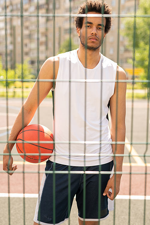 Young serious active player with ball looking at you through fence of basketball court or playground after game