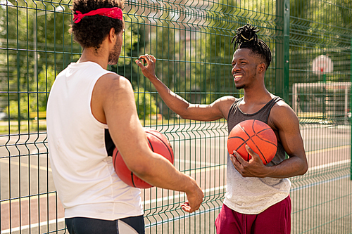 Two young multicultural basketball players interacting after game while standing by fence