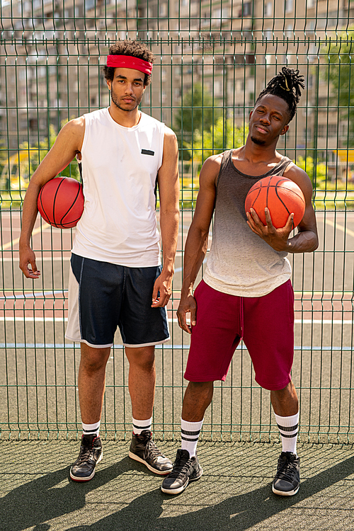 Two young professional multicultural basketball players standing on playground by fence on summer day