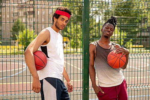 Two active guys in sportswear standing by fence surrounding basketball court while having rest after training