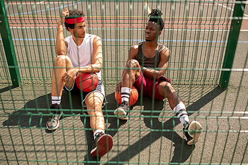 One of young intercultural basketballers telling his friend or playmate about details of game at break by fence