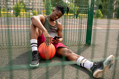 Young African basketball player with smartphone relaxing by fence of playground and scrolling in smartphone