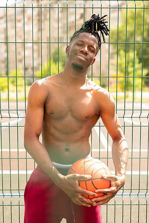 Happy young shirtless basketballer looking at you while standing by fence of playground or court