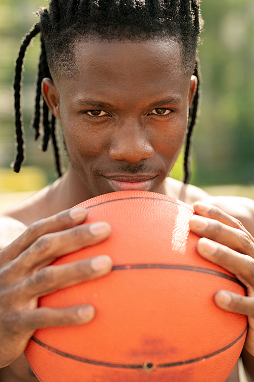 Young successful sports player holding ball for playing basketball by his face while looking at you