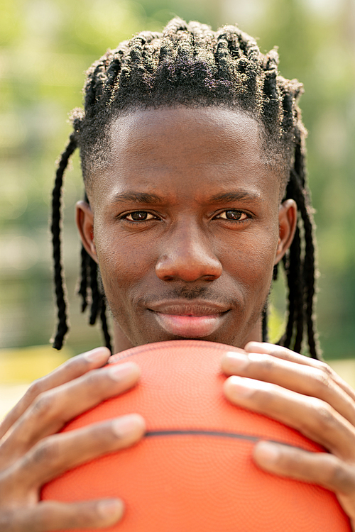Young basketball player holding ball by his face while standing close to camera and looking at you