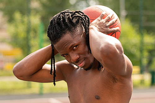 Young successful shirtless basketballer holding ball behind neck while standing in front of camera