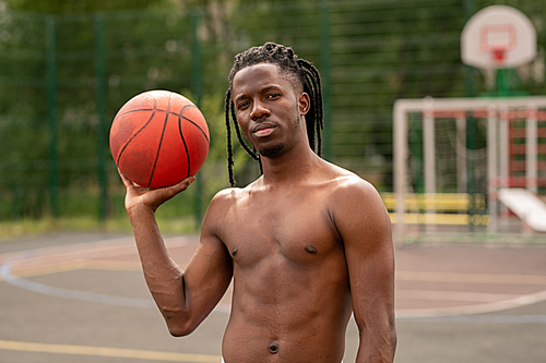 Young shirtless African muscular athlete with ball standing on basketball court in front of camera