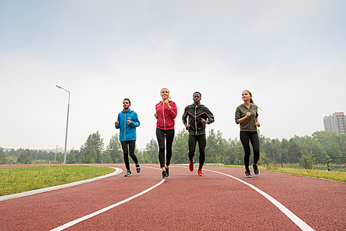 Four young male and female athletes in sportswear running marathon on racetracks of outdoor stadium