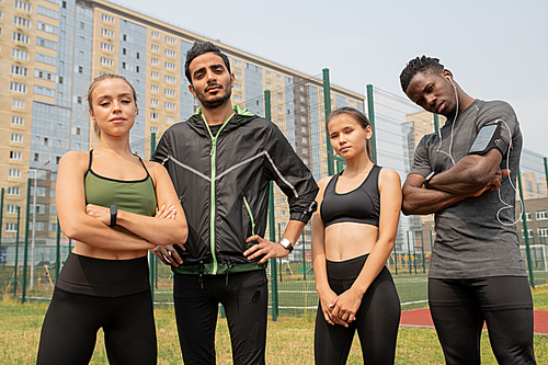 Young sporty men and women in activewear standing in urban environment on outdoor stadium with buildings on background