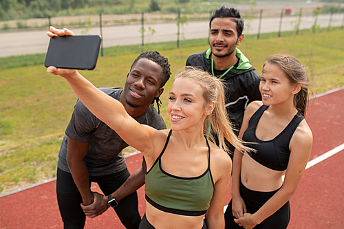 Pretty blonde girl in activewear making selfie with her friends standing near by on outdoor stadium