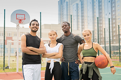 Young cheerful intercultural friends in activewear standing close to one another on basketball playground or stadium