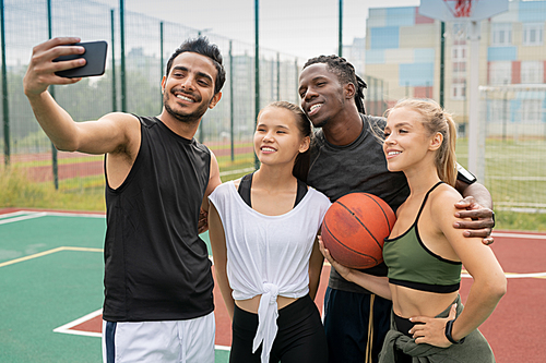 Group of young intercultural friends in sportswear making selfie on outdoor basketball playground or court