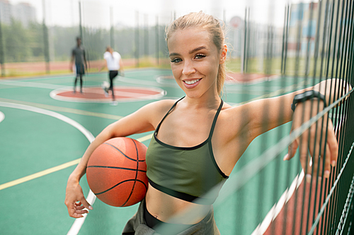 Young successful female basketball player with ball looking at you while standing by net on outdoor playground