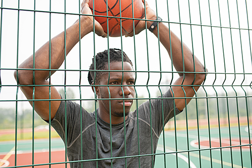 Young basketball player of African ethnicity holding ball over his head while standing in front of sports net