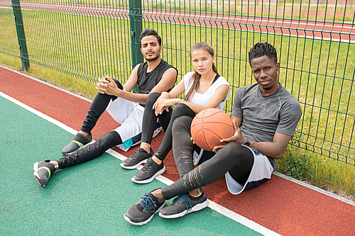 Three young athletes in sportswear sitting on outdoor basketball court by net and having rest after game