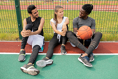 Two guys and girl in sportswear chatting while sitting on playground or basketball court by net
