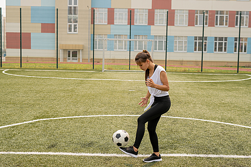Young sportswoman in activewear kicking soccer ball while training on the field or playground before match