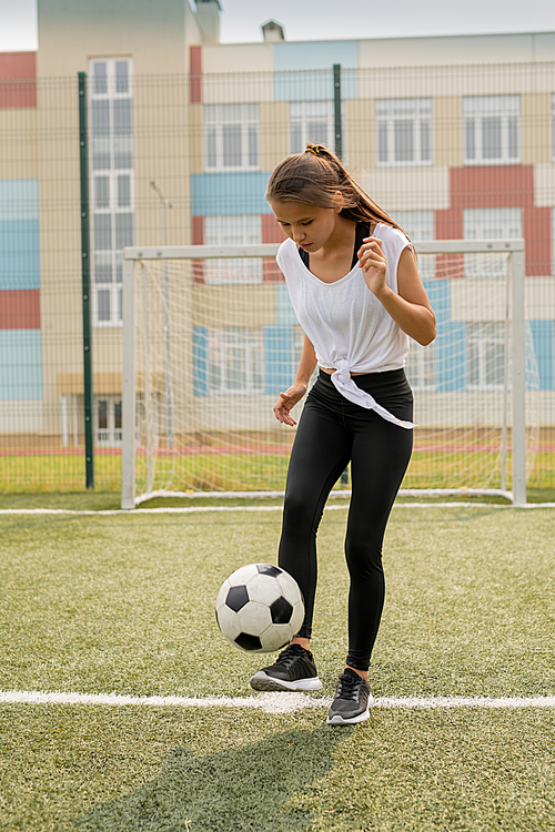 fit girl in sportswear standing on sports field while kicking soccer ball during training in urban