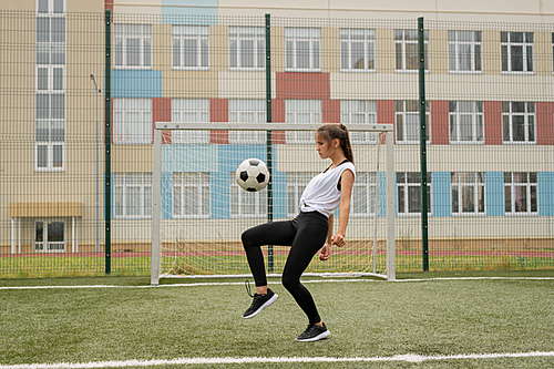 Young sportswoman in leggins, sneakers and t-shirt training to play soccer while kicking ball on outdoor playground