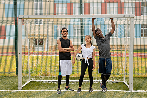 Three young soccer players in sportswear standing by gate on the field in front of camera while having break