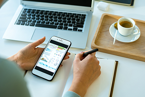 Hands of young contemporary female economist analyzing data on smartphone screen while making notes in notebook