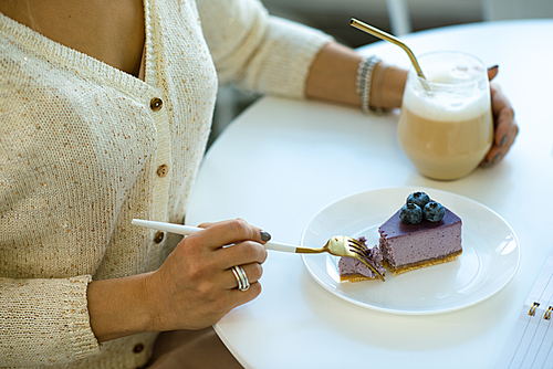 Hands of young woman having cappuccino and eating tasty blueberry cheesecake while sitting by table in cafe