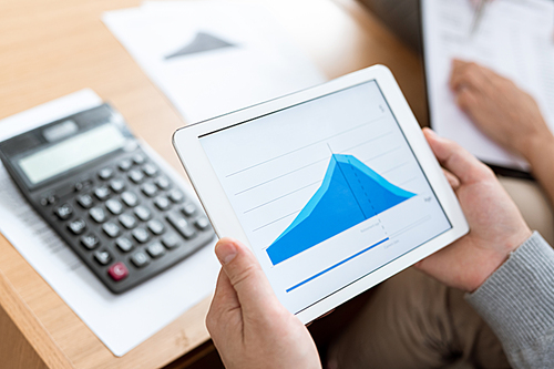 Human hands holding touchpad with blue graph of financial rate on display over calculator on desk