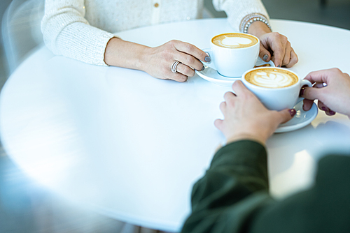 Hands of two young friendly women sitting by table in cafe while having cappuccino during break