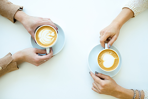 Top view of hands of two friendly females holding cups with cappuccino while relaxing by table in cafe