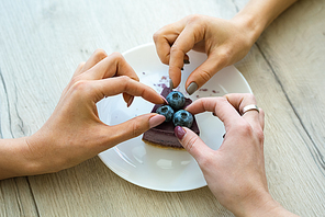 Hands of two girls taking blueberries from top of tasty cheesecake on plate while eating one cake together in cafe