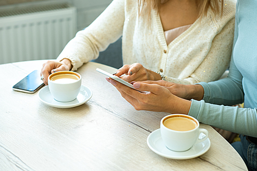 One of two young contemporary women pointing at smartphone screen while discussing online goods with friend by cup of coffee in cafe