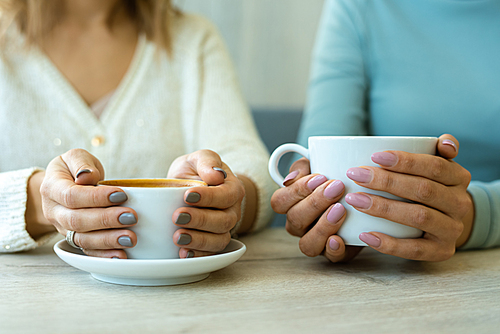 Hands of two young friendly females in casualwear holding cups with coffee while sitting by table in cafe