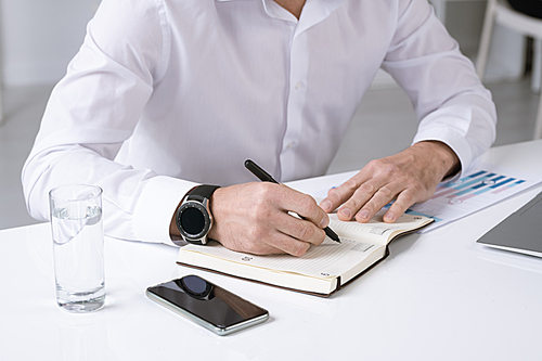Hands of successful male entrepreneur in white shirt making working notes or plan for the day while sitting by desk