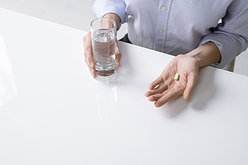 Young sick female or businesswoman in shirt holding glass of water and pill while sitting by desk