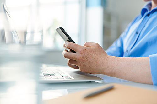 Hands of young contemporary student or employee holding smartphone over workplace while texting