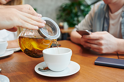 Hands of guy with teapot pouring herbal tea into white porcelain cup while spending time in cafe with college friends