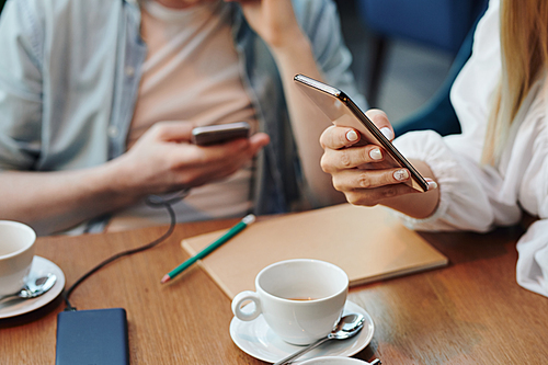 Hand of young mobile woman with smartphone scrolling through online data or contacts over table while having cup of tea