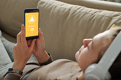 Young learner of online yoga course lying on couch at home and waiting for website download on smartphone screen