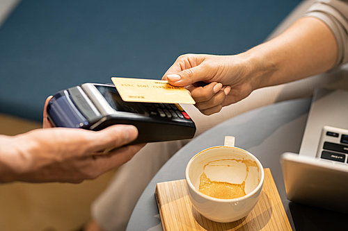 Female hand holding plastic card over electronic payment machine held by waiter while paying for cappuccino in cafe