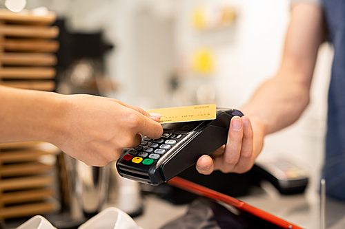 Young client of cafe holding plastic card over electronic payment machine held by waiter or barista while paying for food or drink