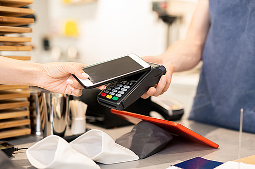 Hand of young woman holding smartphone close to electronic payment machine while paying for food in cafe