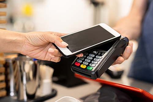 Young woman using her mobile phone for contactless payment while holding it over electronic machine held by barista