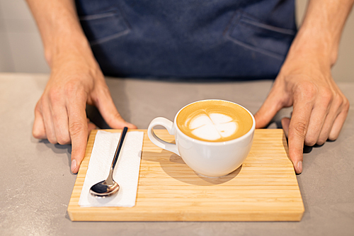 White porcelain cup of cappuccino and small spoon with paper napkin on wooden tray held by young waiter or barista