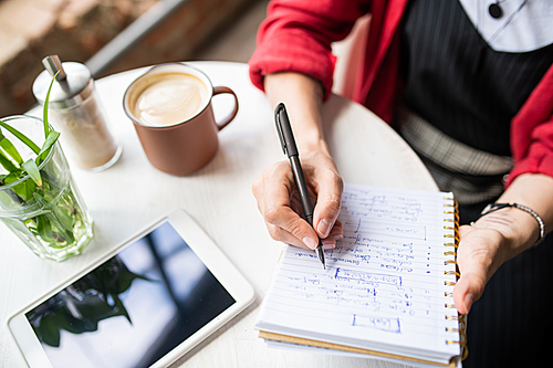 Young woman writing down working notes in notebook while sitting by table in cafe at coffee break