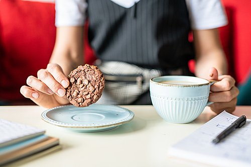 Businesswoman holding round chocolate cookie with nuts over plate while sitting by desk and having tea